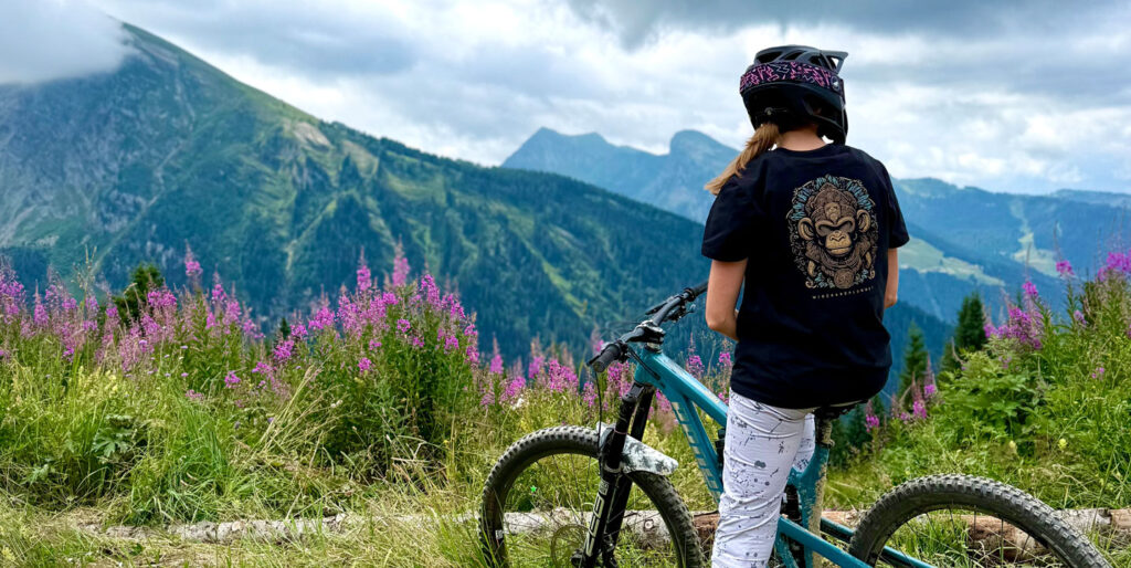 Rider in a Winch & Plummet dritec Ape t-shirt overlooking alpine peaks and wildflowers